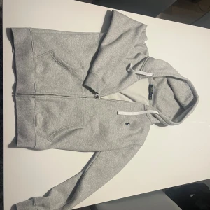  Ralph Lauren zip hoodie - Snygg grå hoodie från Ralph Lauren med klassisk broderad logga på bröstet. Tröjan har huva med vita snören, dragkedja framtill och två stora fickor. Mjuk insida och ribbade muddar vid ärmslut och nederkant. Perfekt för chill dagar.