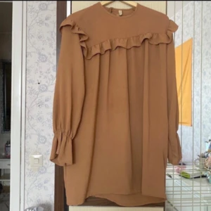 Beige volangklänning från Tablo - Supersöt beige kortklänning från Tablo med volangdetalj över bröstet och långa ärmar med resår och lätt utsvängd avslutning. Klänningen har en rund halsringning och är tillverkad i ett mjukt, strukturerat tyg som känns luftigt och skönt.