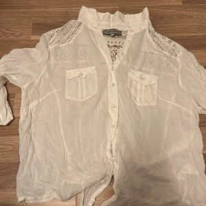 Vit spetsdetaljerad skjorta Lee Cooper - Snygg vit skjorta från Lee Cooper med spetsdetaljer över axlar och rygg. Skjortan har två bröstfickor med lock, knappar framtill och en lite högre krage. Materialet är lätt och luftigt, perfekt för varmare dagar. Spetsen ger en cool och unik touch till looken.