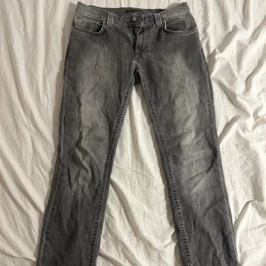 Nudie grå jeans  - Säljer ett par grå jeans från Nudie med klassisk femficksdesign och raka ben. Jeansen har en snygg tvättad look och subtila slitningar. Passar dig som gillar stilrena och tidlösa jeans med bekväm passform.