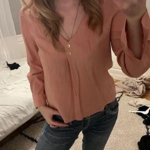 Ljusrosa ZARA blus med v-ringning - Säljer en ljusrosa blus med v-ringad hals och två dekorativa knappar framtill. Blusen har långa ärmar och är tillverkad i ett mjukt, lätt material som känns skönt mot huden. Perfekt att styla med jeans eller kjol för en chill look.