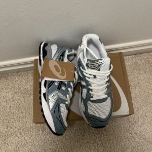 ASICS gel - Snygga ASICS sneakers i vitt och grått med svarta detaljer. Ovandelen är i mesh och syntet för en sportig vibe och bra andningsförmåga. Klassisk snörning och rund tå. Perfekta för dig som gillar stilrena och bekväma skor med retro känsla.