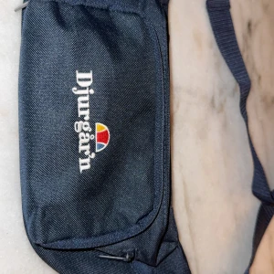 Blå Djurgår'n axelväska/magväska med brodyr - Snygg marinblå axelväska från Djurgår'n med broderad logga och färgglad detalj framtill. Väskan har justerbar rem och praktiska dragkedjor. Perfekt för att bära det viktigaste och visa vilket lag du håller på.