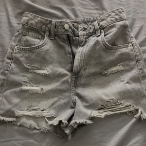 Grå slitna jeansshorts från H&M Divided - Grå jeansshorts från H&M Divided i storlek 34. Råa och fransiga kanter samt flera slitningar framtill för en cool, edgy vibe. Klassisk femficksmodell med knapp och dragkedja. Materialet är en mix av bomull och polyester.