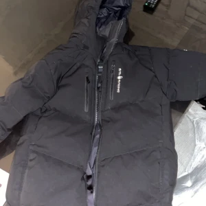 Sail racing jacka  - Svart pufferjacka från Sail Racing med huva och två vertikala bröstfickor med dragkedja. Jackan har en stilren look med logga på bröstet och är perfekt för kalla dagar. Fylld med dun och har lång ärm samt hel dragkedja framtill.