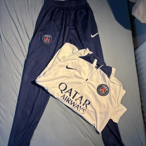 PSG vit och marinblå fotbollströja Nike - Paris Saint-Germain fotbollströja från Nike i vit och marinblå färg med klubbmärke på bröstet och Qatar Airways tryck. Tröjan har korta ärmar och dragkedja vid halsen. Perfekt för dig som vill representera PSG på planen eller till träning.