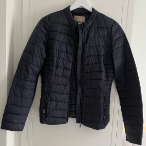 Mörkblå pufferjacka från Orsay - Snygg mörkblå pufferjacka från Orsay med quiltad design och raka linjer. Jackan har ståkrage, långa ärmar och två fickor med dragkedja. Perfekt för kyliga dagar och enkel att matcha med din stil. Dragkedja framtill för smidig stängning. Liten i storleken. Passar bra till vår och sommar då den är en tunnare jacka.