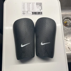 Nike J Guard benskydd XS svart - Säljer ett par svarta Nike J Guard benskydd i storlek XS. De har en stilren design med diagonala linjer och en vit Nike-logga på framsidan. Kommer med original meshpåse och förpackning. Perfekt för fotbollsspelare som vill ha lätt och smidig skyddsutrustning. Jag hade tagit bort etiketten 