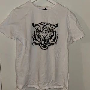 Vit t-shirt med tigertryck Closure London - Vit t-shirt från Closure London i storlek M med ett fett svart tigertryck på bröstet. Klassisk rund hals och korta ärmar. Materialet är mjuk bomull som känns skönt mot huden. Perfekt för dig som gillar djurmotiv och vill sticka ut lite extra.