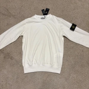 Vit sweatshirt från Stone Island - Vit långärmad sweatshirt från Stone Island med klassisk rund hals och ikonisk svart och gul logotyp-patch på vänster ärm. Tröjan har ribbade muddar och är tillverkad i mjuk bomull för en clean och stilren look.