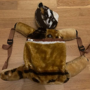 Ryggsäck i plysch med tigerdesign - Ryggsäck i form av en tiger, tillverkad i mjuk plysch med bruna, beige och svarta detaljer. Väskan har justerbara bruna axelremmar och en dragkedja på ryggen.