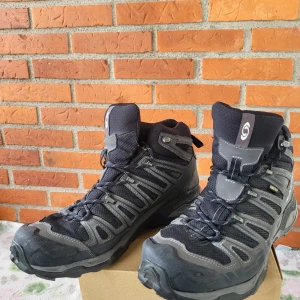 Salomon Gore-Tex kängor svart/grå - Salomon kängor med Gore-Tex, perfekta för äventyr och vandring. Svart och grå färg med robust snörning och slitstark syntet och mesh. Hög modell med grov sula för extra grepp och stabilitet. Salomon-logga på sidan och detaljer i grått.