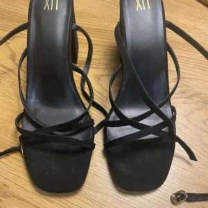 Svarta klackar XIT - Svarta klackar från XIT med smala remmar och fyrkantig tå. Detalj i guld. Materialet är syntet med mockakänsla. Oanvända