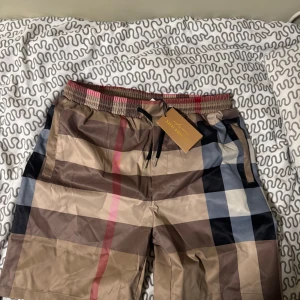 Rutig Burberry shorts  - Säljer ett par sjukt snygga Burberry shorts i klassiskt rutigt mönster med beige, bruna, svarta och röda toner. De har elastisk midja med snörning och är tillverkade i ett lätt syntetmaterial, perfekta för sommaren. Styla med en enkel t-shirt för en clean look. De är strlk XL men ni vet ju hur burberry är de sitter som M