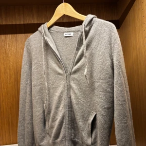 Grå hoodie i kashmir från Soft Goat - Mjuk och lyxig grå hoodie från Soft Goat i storlek M, tillverkad i kashmir. Tröjan har huva med snörning, dragkedja framtill och två fickor. Perfekt för dig som gillar chill och stilren look. Långärmad och ribbad vid muddar och nederkant.