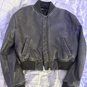 Grå croppad bomberjacka från Zara - Snygg grå bomberjacka från Zara med croppad passform och ribbade muddar. Jackan har dragkedja framtill med cool snörningsdetalj och två fickor. Materialet är syntet med skinnlook, perfekt för dig som gillar streetstyle.