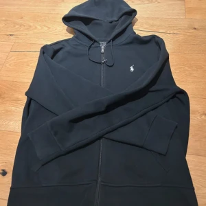 Svart hoodie från Polo Ralph Lauren - Svart hoodie från Polo Ralph Lauren med dragkedja och huva. Klassisk design med vit broderad logga på bröstet och snörning vid huvan. Tillverkad i mjuk bomull, perfekt för chill dagar. Långärmad och med fickor framtill.