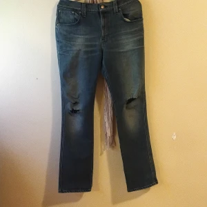 Blå Nudie Jeans med slitna knän - Säljer ett par blå jeans från Nudie Jeans med klassisk femficksdesign och slitna detaljer vid knäna. Jeansen har raka ben och subtila slitningar framtill, samt Nudie Jeans logga på bakfickan. Materialet är robust denim i bomull.
