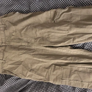 Beige cargopants från STR, strl 38 - Säljer ett par beige cargopants från STR i storlek 38. Byxorna har flera praktiska fickor på sidorna och bak, samt bälteshällor. Materialet är mjukt och slitstarkt, perfekt för dig som gillar en avslappnad och funktionell stil.