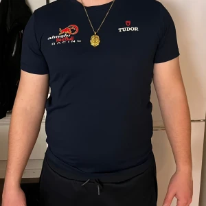 Sail racing Alinghi Red Bull Racing t-shirt marinblå - Marinblå t-shirt från Sail Racing x Alinghi Red Bull Racing med tryckta loggor på bröstet och ärmarna. Klassisk rund hals och korta ärmar. Materialet är mjukt och stretchigt, perfekt för dig som gillar sportig stil och racing-vibe.