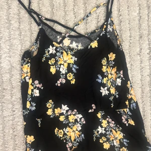 Svart blommig kortklänning H&M - Supersöt svart kortklänning från H&M med tunna axelband som korsas i ryggen. Klänningen har ett färgglatt blommönster i gult, vitt, blått och rosa och är gjord i ett lätt och luftigt material. Perfekt för varma dagar när du vill känna dig extra fin.