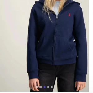 Marinblå hoodie från Polo Ralph Lauren - Snygg marinblå hoodie från Polo Ralph Lauren med röd logga på bröstet. Tröjan har huva, dragkedja och två praktiska fickor framtill. Perfekt för dig som gillar klassisk streetstyle och vill ha en bekväm och stilren look.