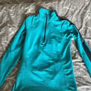 Turkos Nike Dri-Fit träningströja XS - Nike Dri-Fit långärmad träningströja i turkos med svart detalj längs ärmarna och half zip framtill. Snyggt Nike-logga på bröstet och hög krage. Perfekt för löpning eller gym. Materialet är polyester som andas och transporterar bort fukt.