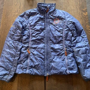 Blå pufferjacka från Chriwen, S - Snygg blå pufferjacka från Chriwen med broderad logga på bröstet och ryggen. Jackan har hög krage, två sidofickor med dragkedja och detaljer i brunt läder på dragkedjorna.  NYPRIS 2500