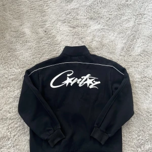 Svart ziptröja Corteiz med vit logga - Svart ziptröja från Corteiz i storlek Large med vit logga tryckt både fram och stort på ryggen. Tröjan har vita piping-detaljer längs ärmarna, ribbade muddar och hel dragkedja. Perfekt för dig som gillar streetwear och vill sticka ut med snygg branding.