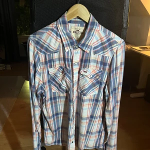 Rutig skjorta från Hollister XL - Snygg rutig skjorta från Hollister i storlek XL. Skjortan har blå, röda och beige rutor, klassisk krage och två bröstfickor med knappstängning. Tillverkad i mjuk bomull, perfekt för en chill och avslappnad stil. Knappar framtill och på fickorna ger extra detaljer.