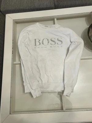 Vit sweatshirt från Hugo Boss - Säljer en stilren vit sweatshirt från Hugo Boss med stort silverfärgat BOSS-tryck på bröstet. Tröjan har rund halsringning, långa ärmar och ribbade muddar. Perfekt för dig som gillar klassisk och enkel design med en exklusiv touch.
