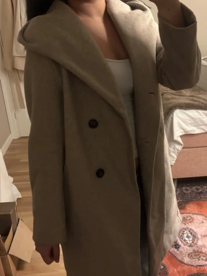 Kappa - Säljer en beige kappa med breda slag och dubbelknäppning med svarta knappar. Jackan har en rak och loose passform, är lång och har klassisk design. Materialet känns mjukt och har ull-look, perfekt för dig som gillar stilrena och enkla plagg. (inget skärp tillkommer)