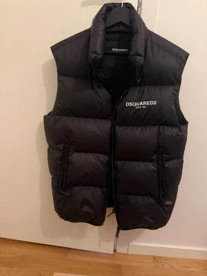 Svart dunväst från Dsquared2 - Säljer en svart dunväst från Dsquared2 med hög krage och två dragkedjeförsedda fickor. Västen har en clean look med Dsquared2-logga på bröstet och stängs med dragkedja framtill. Perfekt för lager-på-lager under kyliga dagar.