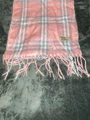 Rutig rosa halsduk från Burberry - Säljer en klassisk halsduk från Burberry i rosa med rutigt mönster i vitt och grått. Halsduken har fransar i ändarna och är tillverkad i mjukt material som känns lyxigt mot huden. Perfekt accessoar för att lyfta din outfit.