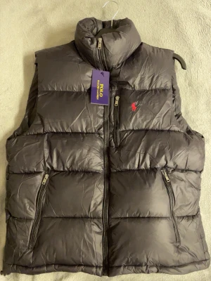 Svart dunväst från Polo Ralph Lauren - Snygg svart dunväst från Polo Ralph Lauren med hög krage och dragkedja framtill. Västen har två snedställda fickor med dragkedja och den klassiska röda loggan broderad på bröstet. Perfekt för lager-på-lager och riktigt chill stil.