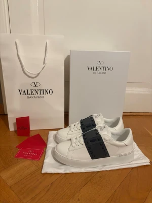Valentino Garavani Rockstud sneakers - Säljer ett par vita Valentino Garavani Rockstud sneakers i skinn med svart band och ikoniska nitar över vristen. Klassisk rund tå, snörning och platt sula med mönstrad undersida. Kommer med originalkartong, påse, valentino kort och dustbag. Tveka inte att fråga om ni har någon fråga😀