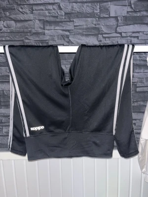Svarta träningsbyxor med vita ränder - Säljer ett par klassiska svarta träningsbyxor från Adidas med vita ränder längs sidorna. Byxorna har en normal passform och är tillverkade i ett mjukt syntetmaterial som är perfekt för träning. Adidas-loggan syns på ena benet.