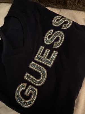 Mörkblå tröja med strass från Guess - Snygg mörkblå långärmad tröja från Guess med v-ringning och stora glittriga strassbokstäver över bröstet. Tröjan är i mjukt material och har en slim passform som sitter snyggt på kroppen. Perfekt för dig som vill sticka ut lite extra.