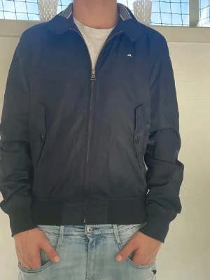 Marinblå bomberjacka från J.Lindeberg - Säljer en stilren marinblå bomberjacka från J.Lindeberg. Jackan har dragkedja framtill, ribbade muddar och krage med randig detalj. Två fickor med knappstängning och diskret logga på bröstet. Perfekt till jeans och t-shirt. Mitt pris 449 men går att diskutera. Storlek L men går med M också
