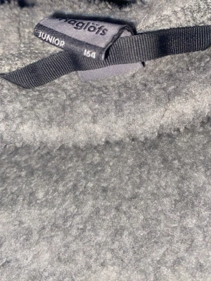 Grå fleecehoodie från Haglöfs Junior - Mysig grå hoodie från Haglöfs Junior i storlek 164. Tillverkad i mjuk fleece med huva och broderad logga på bröstet. Perfekt för kyliga dagar och outdoor-häng. Snygg och enkel design som passar till det mesta. Som ni ser på bilden är den lite sönder vid drag kedjan men de är inget som påverkar kedjan