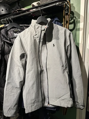 *helt ny* vind/skidjacka från Ralph Lauren - Snygg grå vindjacka från Polo Ralph Lauren med klassisk logga på bröstet. hög krage för halsskydd. Helt nya oanvänd. köpt för 1700 kr