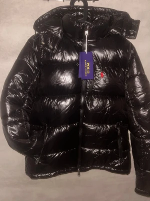 Svart pufferjacka från Polo Ralph Lauren - Säljer en svart pufferjacka från Polo Ralph Lauren med blank finish och röd broderad logga på bröstet. Jackan har dragkedja framtill, hög krage och praktiska fickor med dragkedja. Perfekt för kalla dagar och riktigt snygg till streetwear.