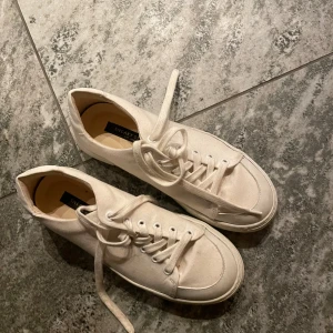 Vita sneakers från Sneaky Steve - Säljer ett par klassiska vita sneakers från Sneaky Steve. Skorna har rund tå, snörning och platt sula. Tillverkade i bomullstyg med stilren design som passar till det mesta. Perfekta för dig som gillar enkel och clean stil.