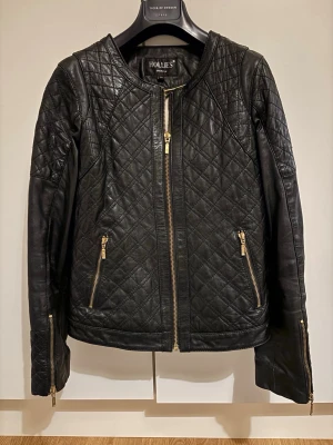 Svart quiltad skinnjacka Hollies - Säljer en svart skinnjacka från Hollies med quiltat mönster och guldfärgade dragkedjor. Jackan har lång ärm, rund halsringning och dragkedjor vid fickor och ärmslut. Snygg och tidlös modell som lyfter varje outfit.