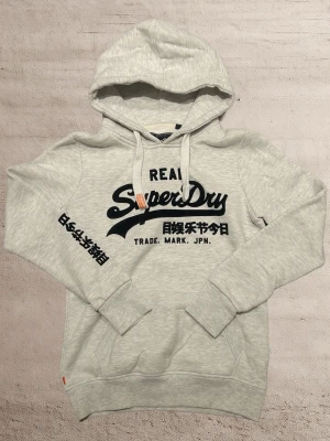 Superdry Hoodie XS - En väldigt snygg hoodie från Superdry i färgen vit/grå med svart tryck. Storlek XS. Mycket bra skick! 😀