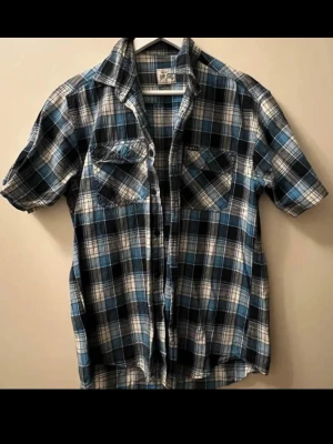 Blårutig kortärmad skjorta - Helt ny, oanvänd kortärmad skjorta med blå, svart och vit rutig design. Två bröstfickor med lock och knappar framtill. Skjortan har klassisk krage och är perfekt för dig som gillar avslappnad stil.