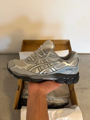  Asics gel nyc grå - Säljer ett par stilrena Asics sneakers i grått med silvriga detaljer och svart sula. Skorna har meshpaneler för bra ventilation och klassisk snörning. Perfekta för dig som gillar sportig och clean stil. Storlek EU 42.