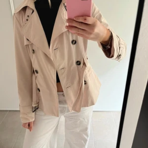 Beige trenchcoat med knappar - Säljer en snygg beige trenchcoat med dubbelknäppning och stora knappar. Jackan har klassisk krage, bälte vid ärmslut och figurnära passform. Perfekt för dig som vill ha en stilren och tidlös jacka till vår eller höst.