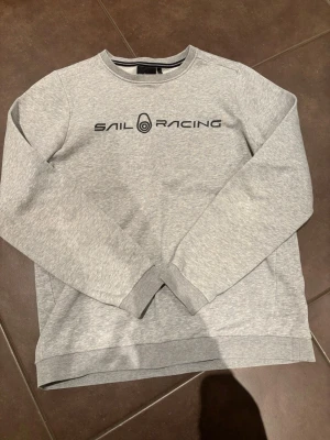 Grå sweatshirt från Sail Racing - Snygg ljusgrå sweatshirt från Sail Racing med svart logga tryckt på bröstet. Klassisk rund halsringning och långa ärmar. Tillverkad i mjuk bomullsmix som är skön att ha på sig. Perfekt till chill dagar eller när du vill ha en clean look.