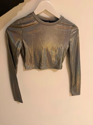 Metallic croppad långärmad topp - Säljer en croppad långärmad topp i metallic silver med glansig finish. Toppen har rund halsringning och är figurnära i modellen. Perfekt för dig som vill sticka ut med en futuristisk vibe.